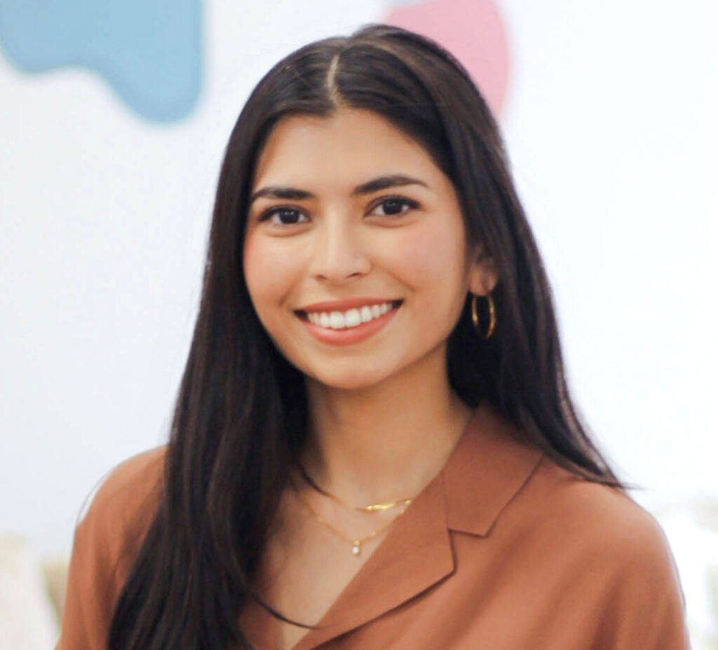 Nuha Siddiqui smiles as she stands in front of a display of colourful abstract shapes.