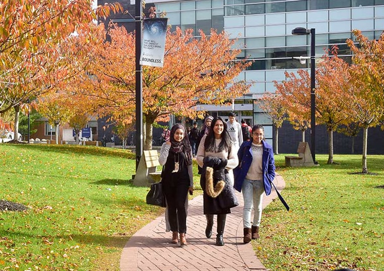 Three smiling students walk in a row along a path at UTSC, under small trees.