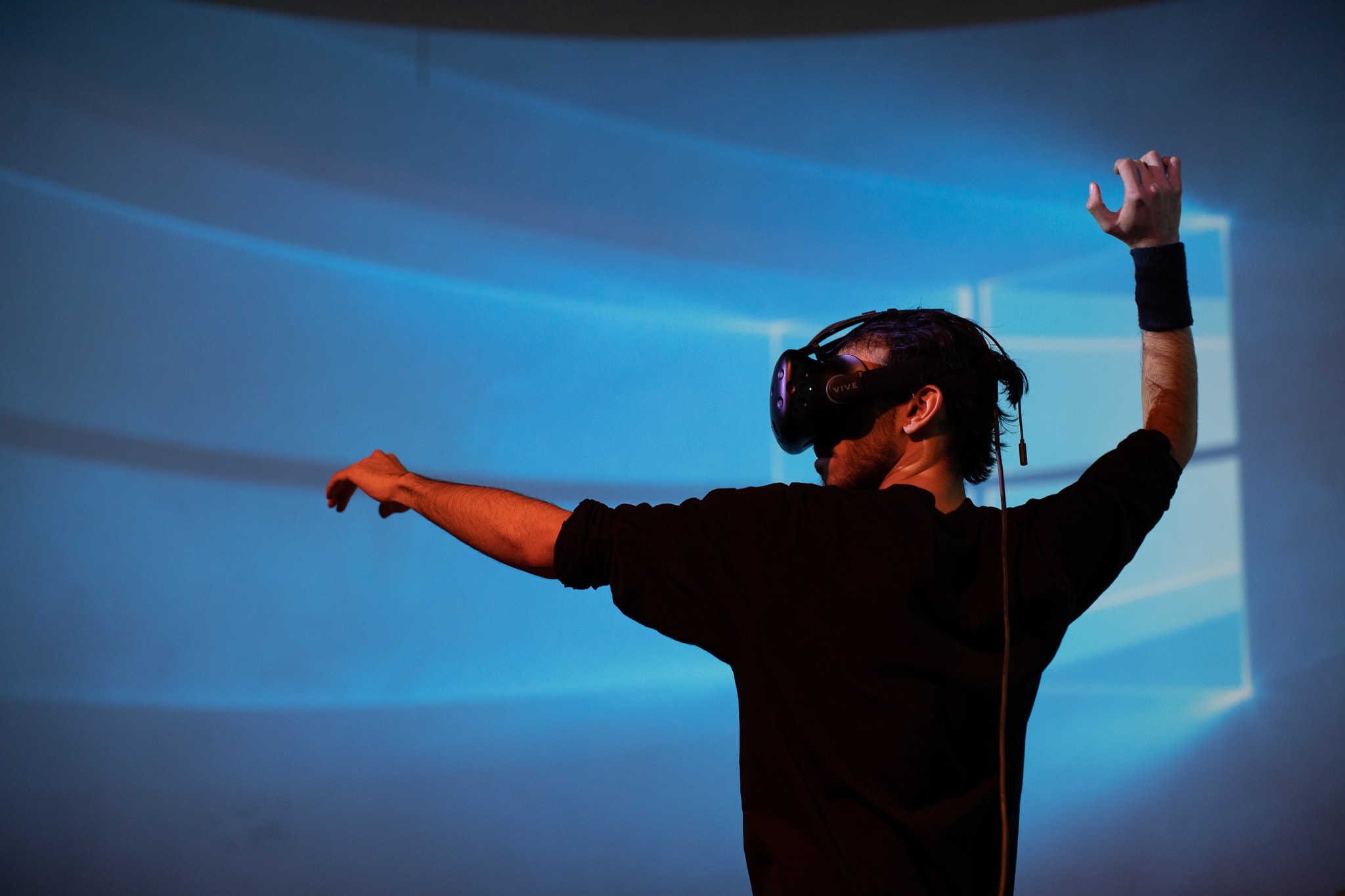 A man wears a virtual reality headset over his eyes. He stretches out one arm and reaches up with the other.