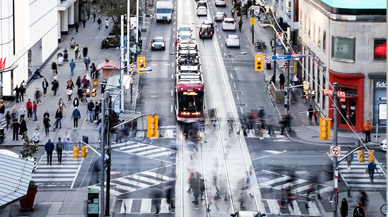 An overhead view of a busy Toronto intersection with cars and a streetcar. People cross the road and chat on the sidewalk.