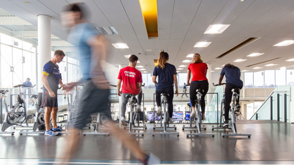 A group of trainers and students pedal stationary bikes while a man jogs on a track in the foreground