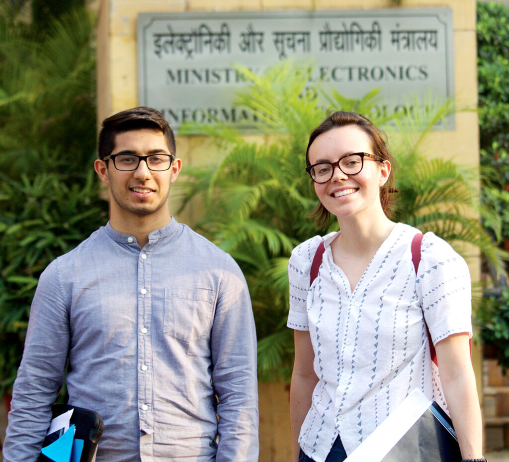 A young man and woman smile in front of a sign that reads, in English and Devanagari alphabets, Ministry of Electronics.