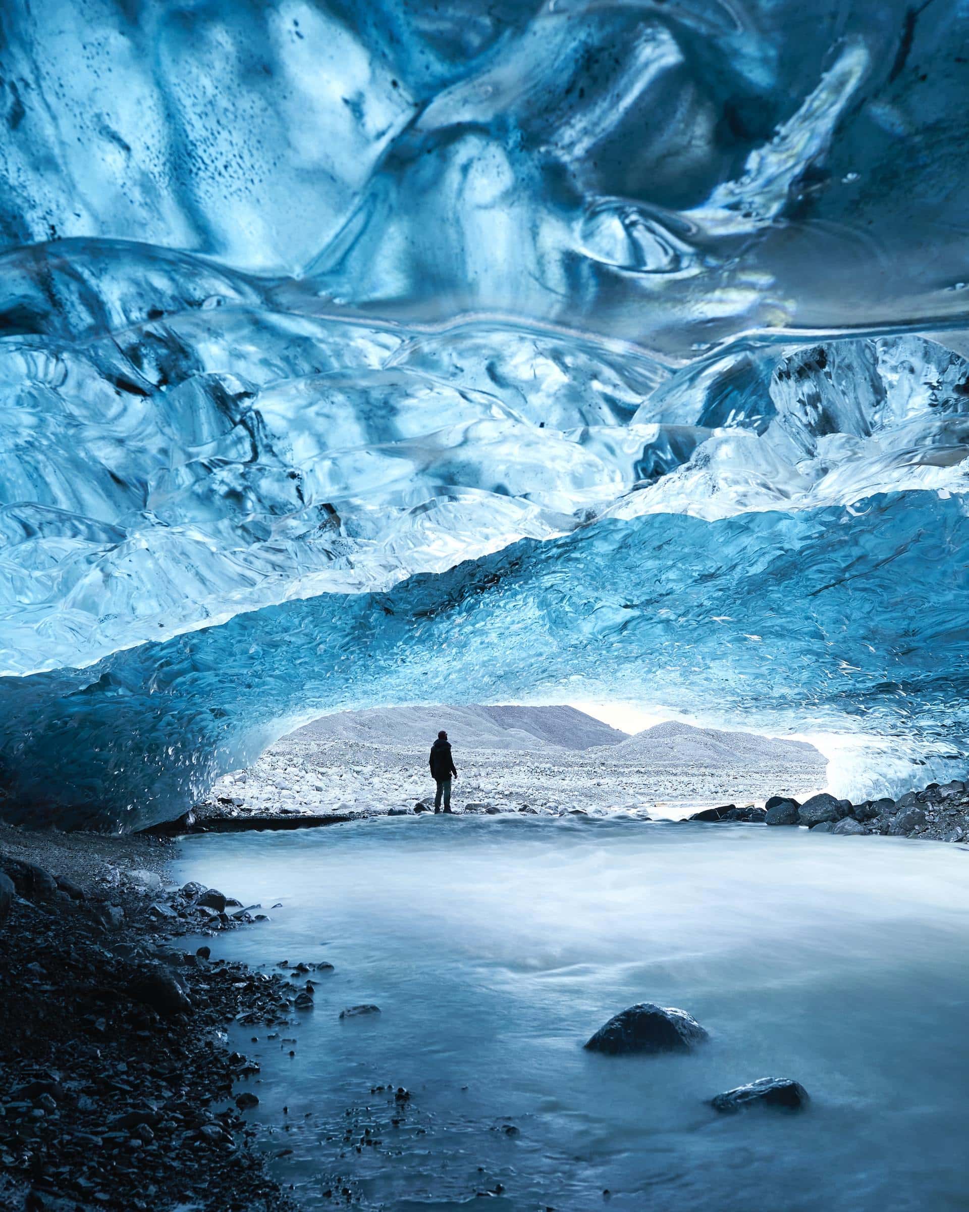 A person stands in the entrance of a huge cave, looking out. The cave ceiling is made of ice with light reflecting through.