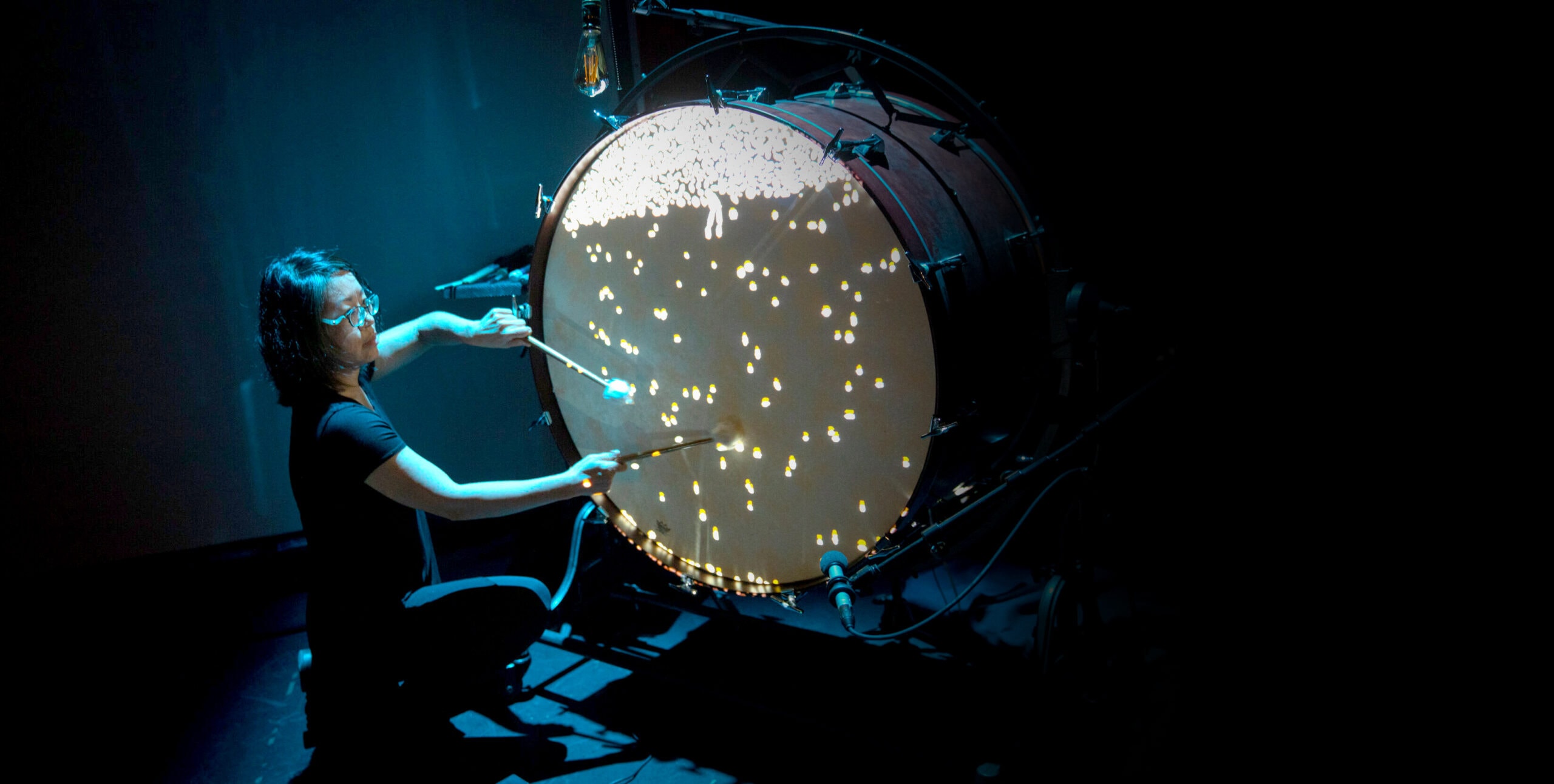 A woman kneels and strikes a metre-high drum. Blobs of light are scattered over the drum surface and cluster at the top.