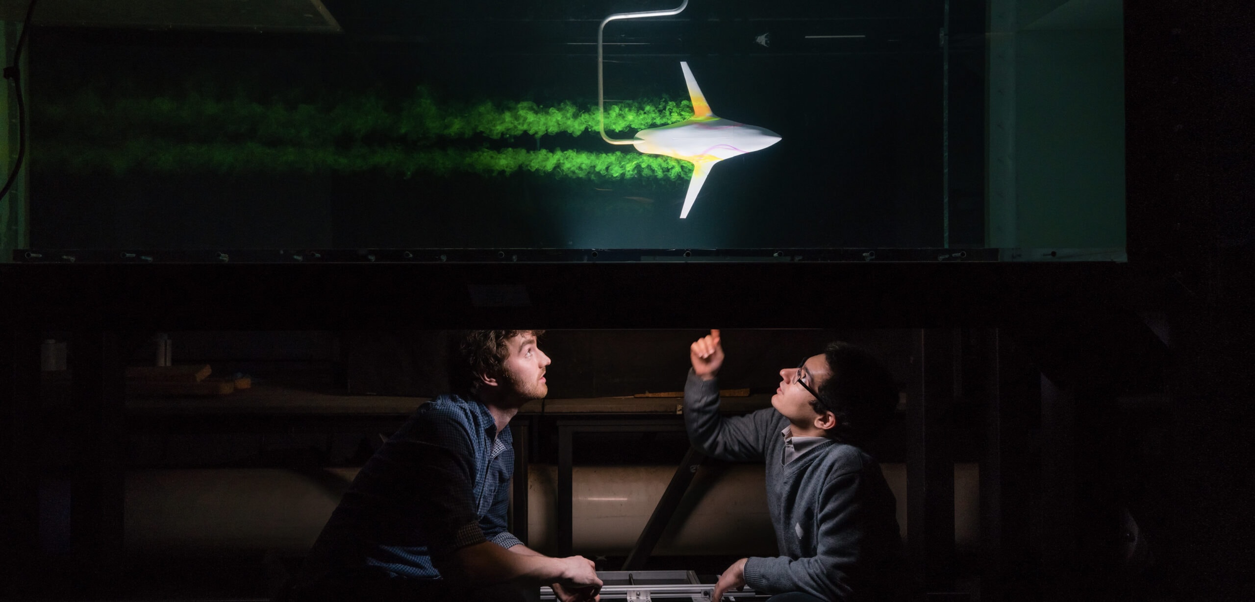In a darkened lab, two men look up at a tank, where an airplane-shaped object leaves bright, turbulent vapour trails.