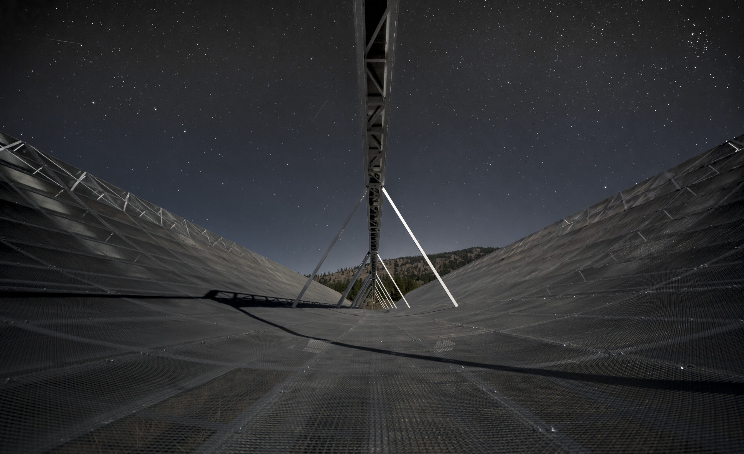 A view from inside the CHIME telescope, showing a grid-like metal floor curving upward. Overhead, the sky is full of stars.