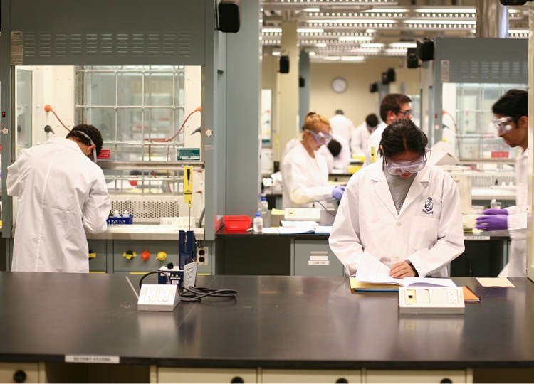 A diverse group of people in lab coats and safety goggles work intently the beakers, books and equipment in a lab at U of T's Department of Chemistry.
