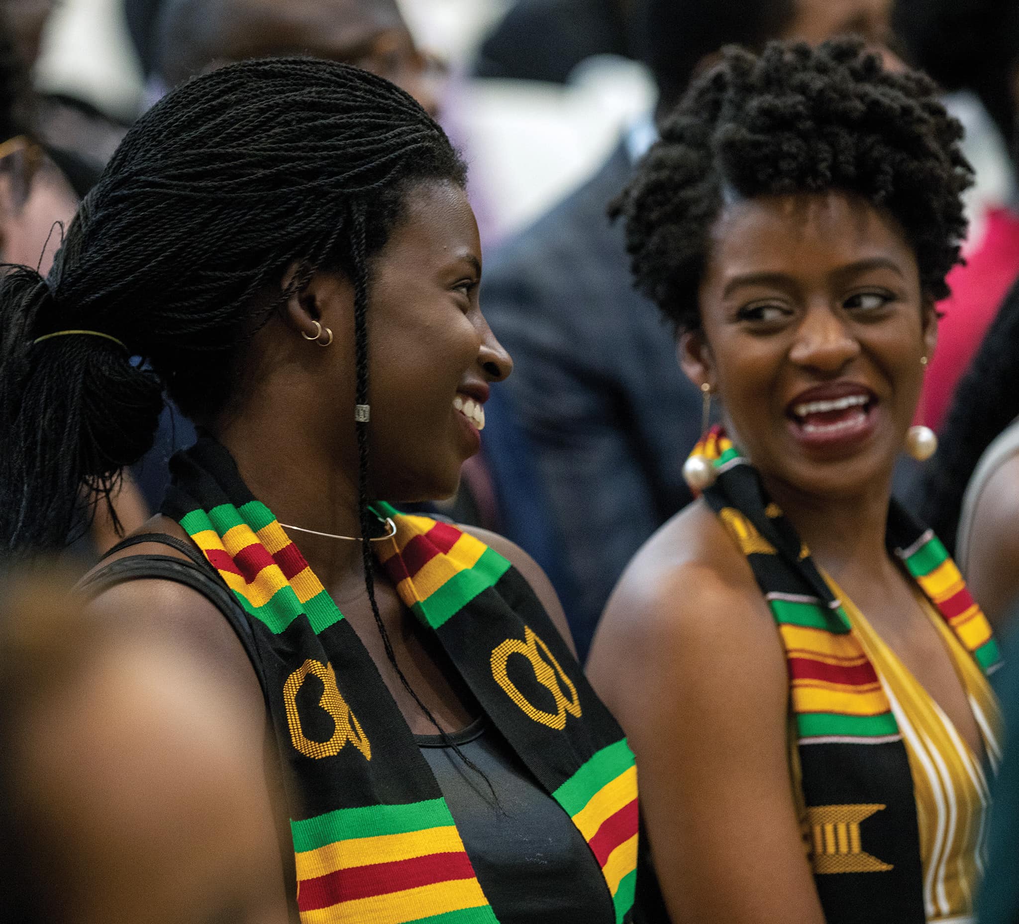 Two Black women laugh happily together. Both are wearing scarves in the pan-African colours of green, yellow and red.