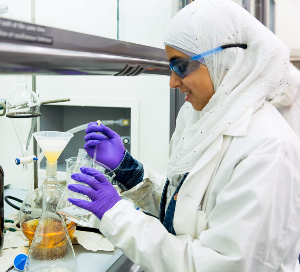 A woman wears a lab coat, gloves, goggles and a hijab. She decants a clear liquid while standing at a bench full of beakers. 