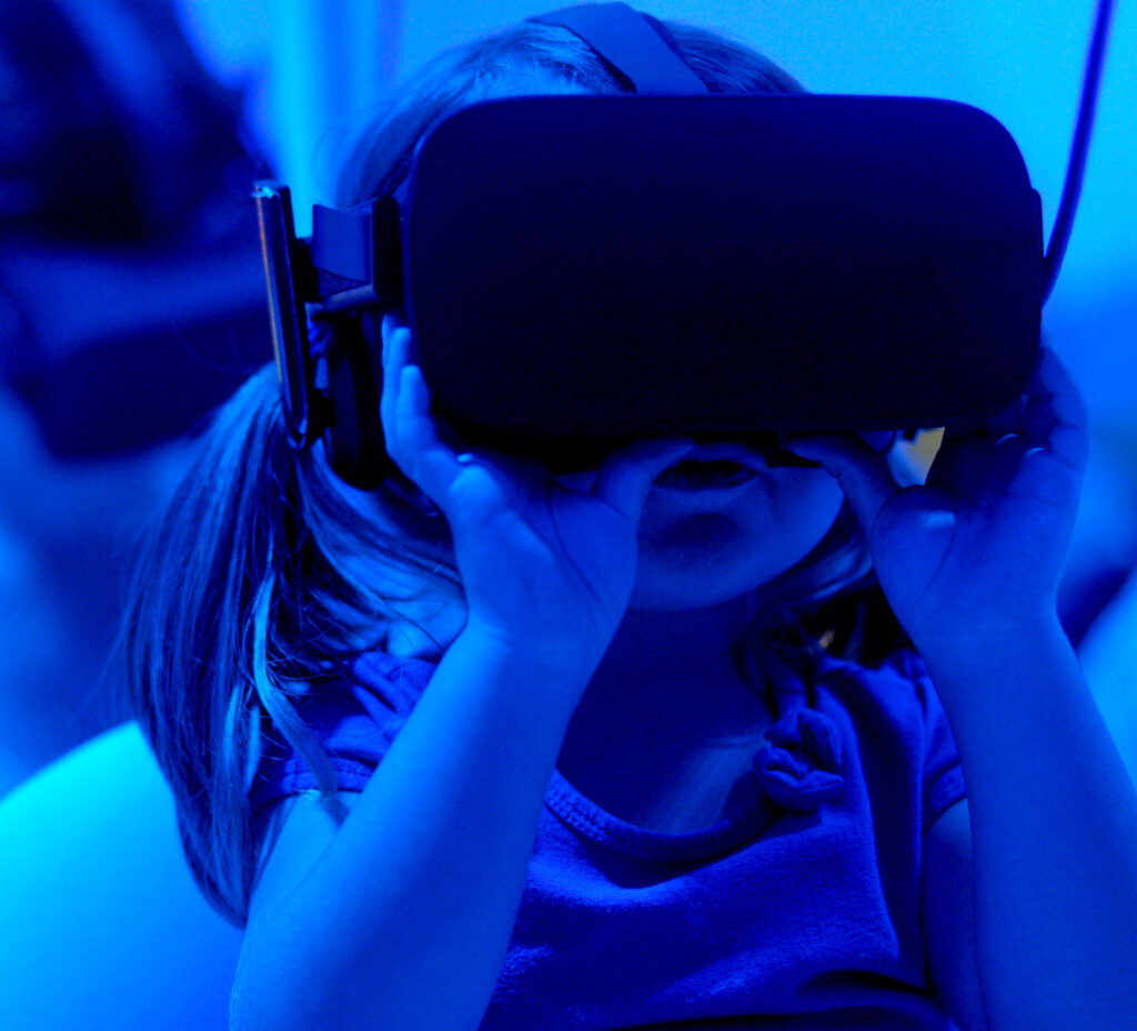 A toddler peers into a virtual reality headset strapped to her head. She is in a room lit with blue light.