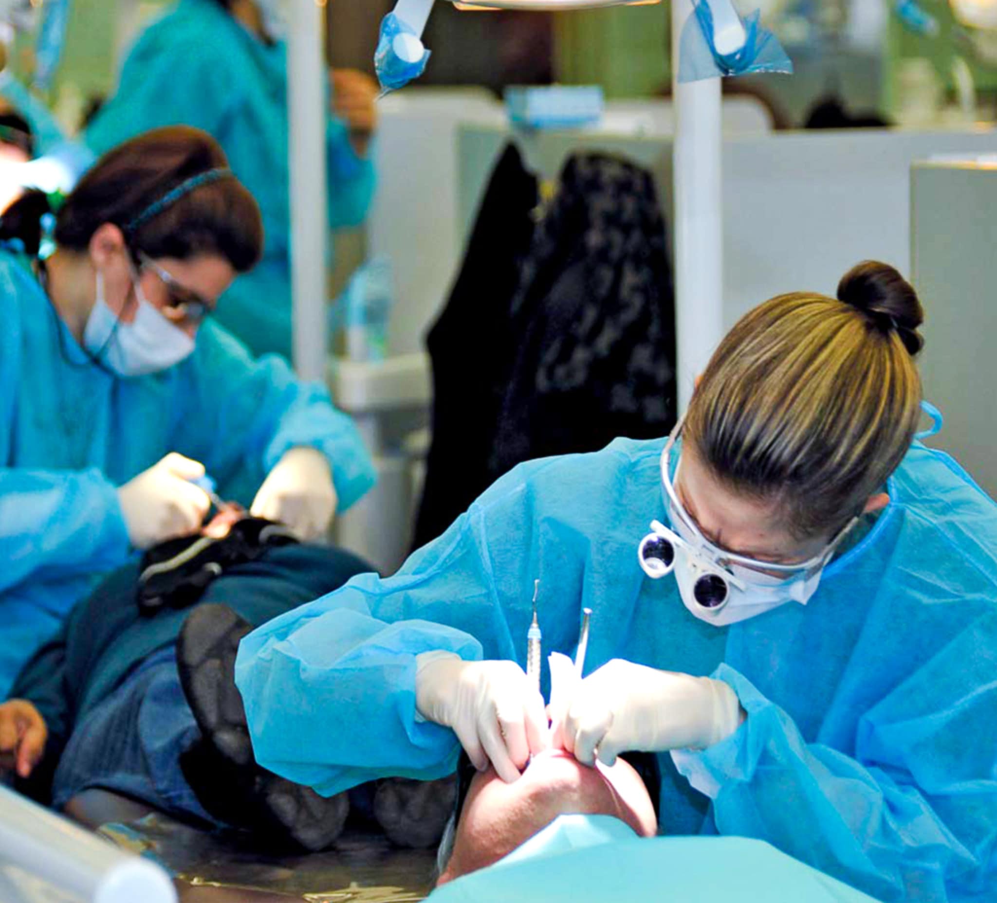 Two women wear goggles, masks, gowns and gloves. They use dentist tools to work on two patients in adjacent cubicles.