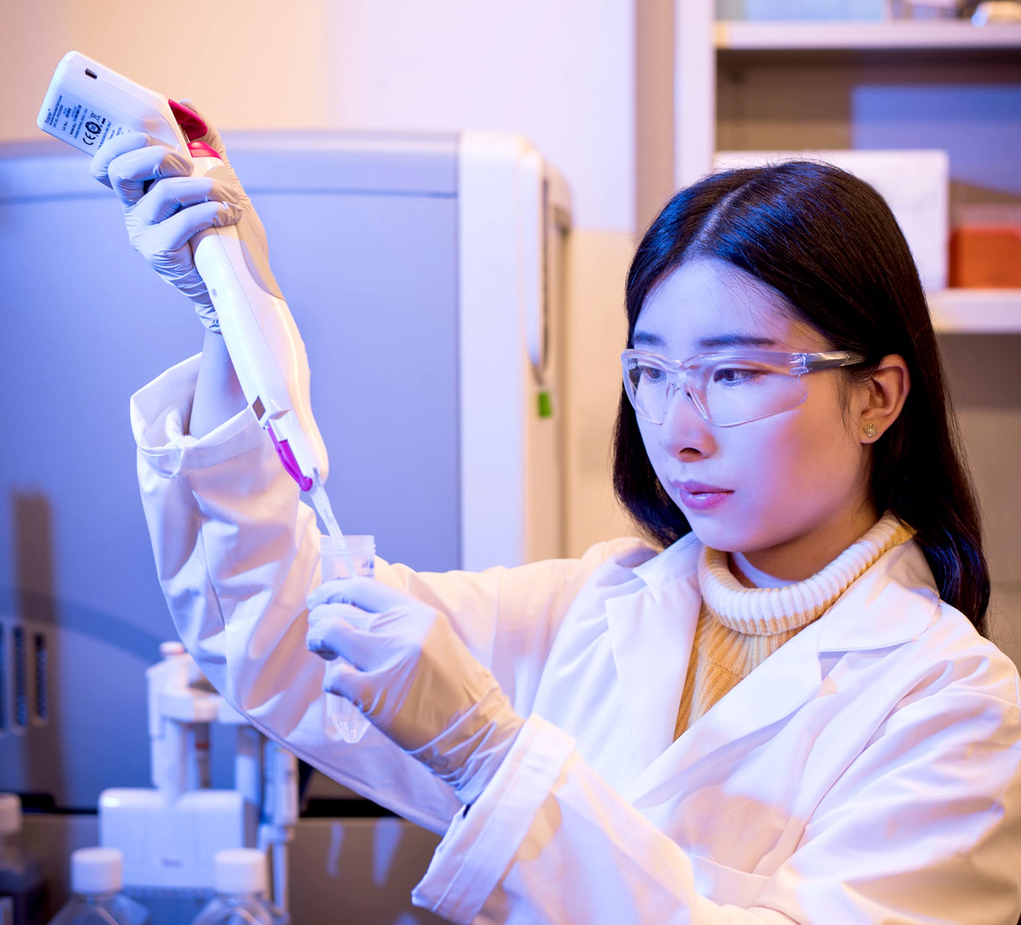 A woman wears a lab coat, gloves and goggles. She uses a handheld gadget to add liquid to a plastic test tube.