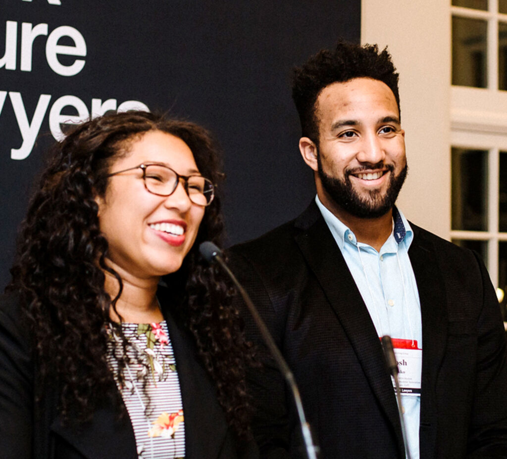 A young Black woman and man smile as they stand at a podium. A sign above them reads: Black Future Lawyers.