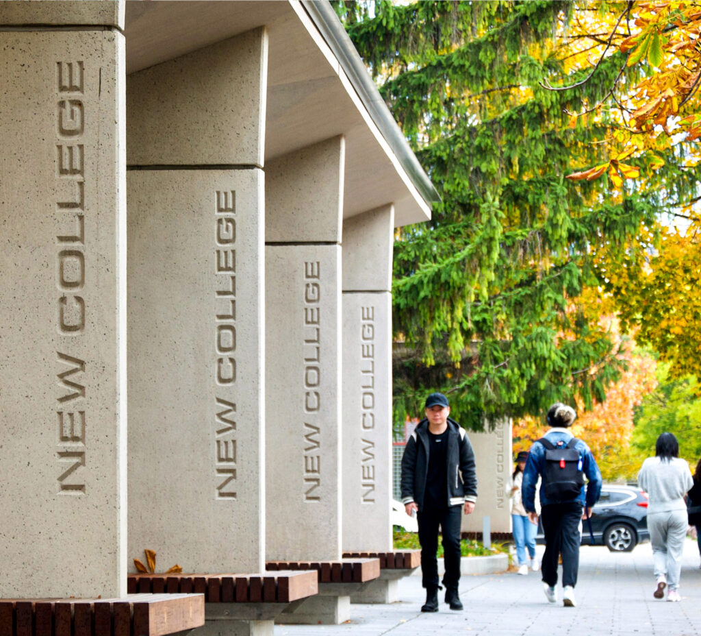 People walk past a concrete shelter, where walls stand between wooden benches. Text carved on the walls reads, New College.