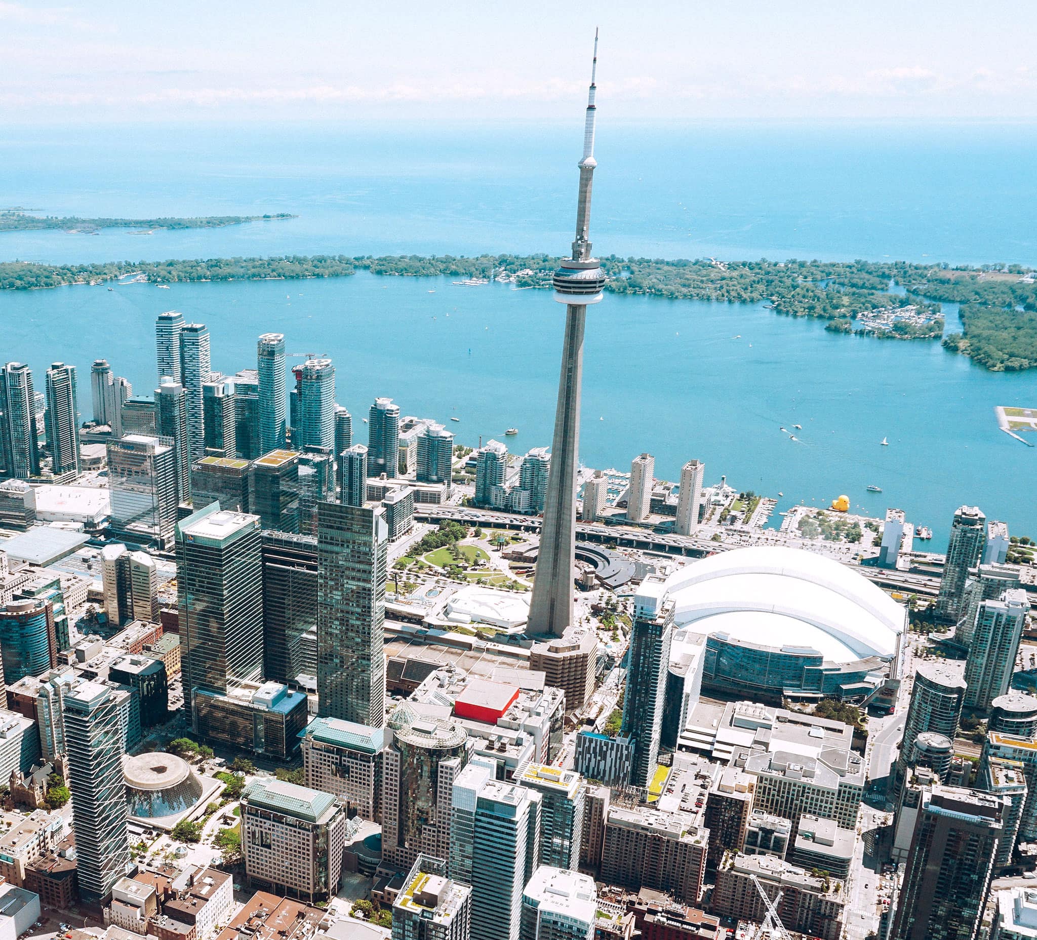 An aerial view of downtown Toronto on a sunny day, showing the CN Tower, Rogers Centre, the harbour and the Toronto islands.