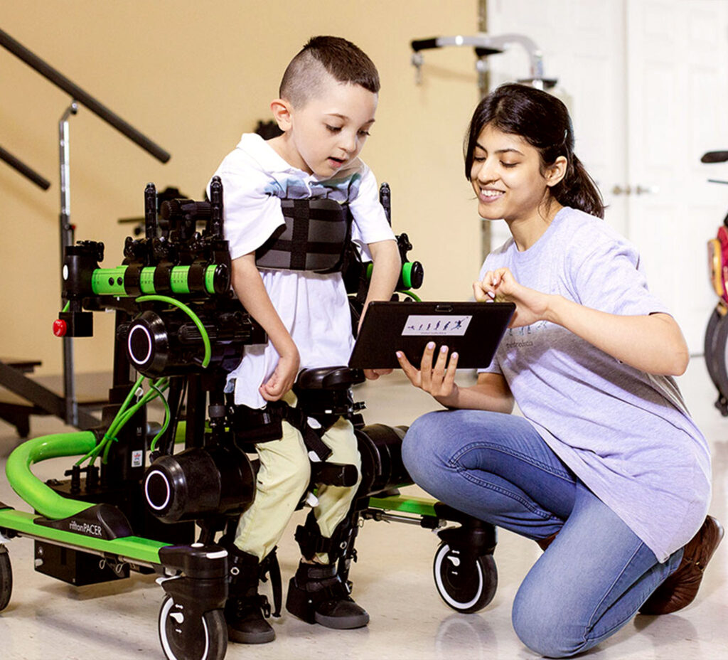 A small, smiling boy is strapped into a wheelchair-like device that holds him upright with his feet touching the ground.