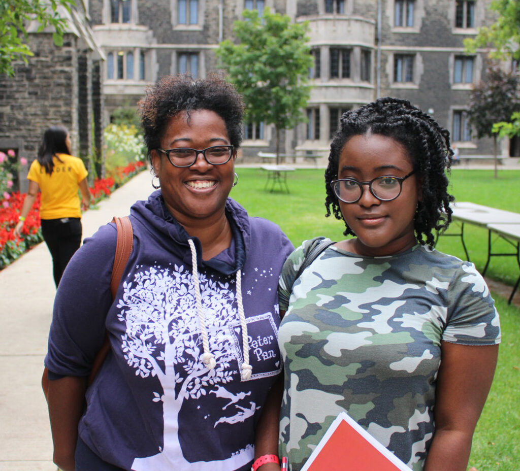 Two women students smile together, standing in a flower-filled quadrangle at Victoria College.