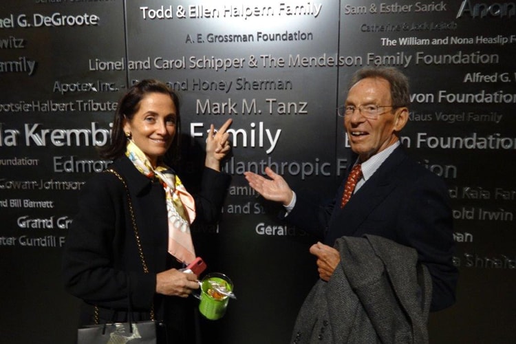Jacqueline and Mark Tanz smile while pointing at Mark Tanz's name on a wall listing donors to the Tanz Centre.