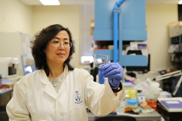 Lidan You, wearing a lab coat, safety glasses and gloves, holds up a small clear square with tiny coloured dots on it.