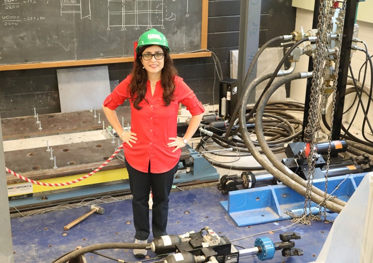 Oya Mercan smiles, standing surrounded by pipes, chains, valves, girders, a hammer and a chalkboard covered with diagrams.