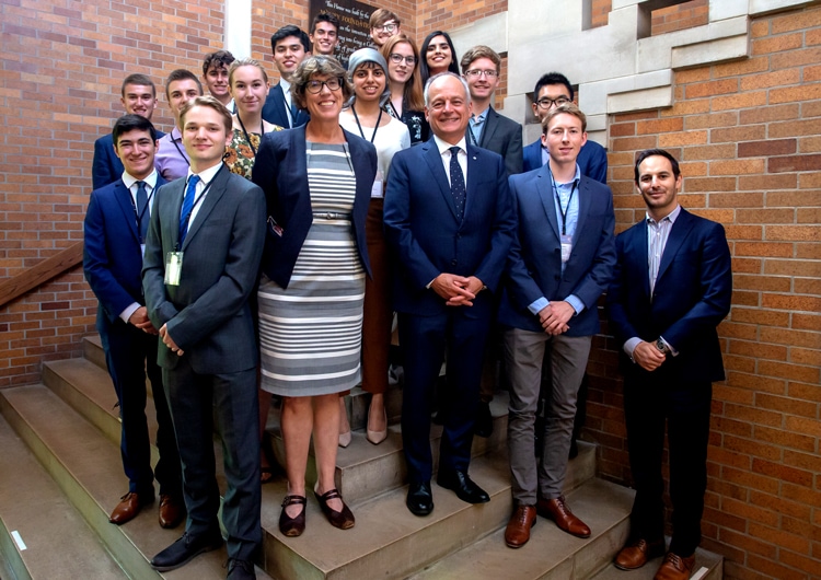 Several students stand smiling on on the stairs outside Massey College with President Meric Gertler