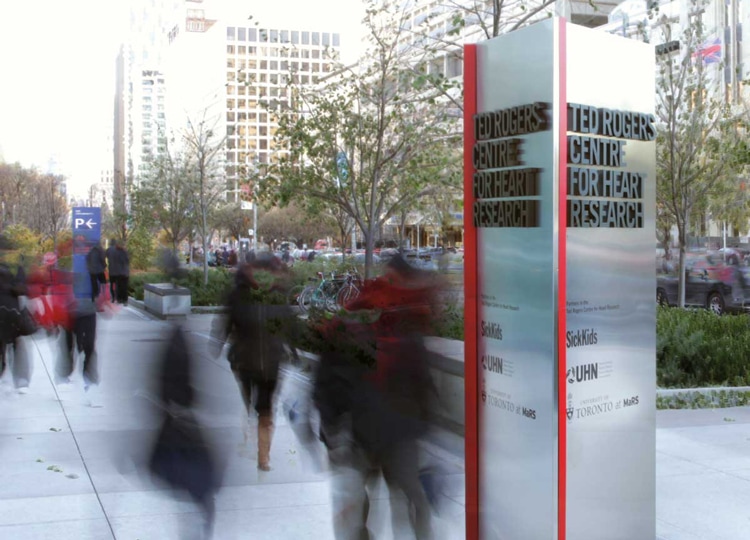 People walk along University Ave in Toronto past a pillar with raised letters reading: Ted Rogers Centre for Heart Research.