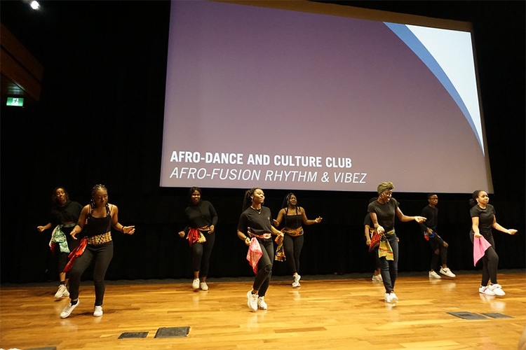 Eight people dance on a stage beneath a banner that says: Afro-Dance and Culture Club, Afro-fusion rhythm and vibez.