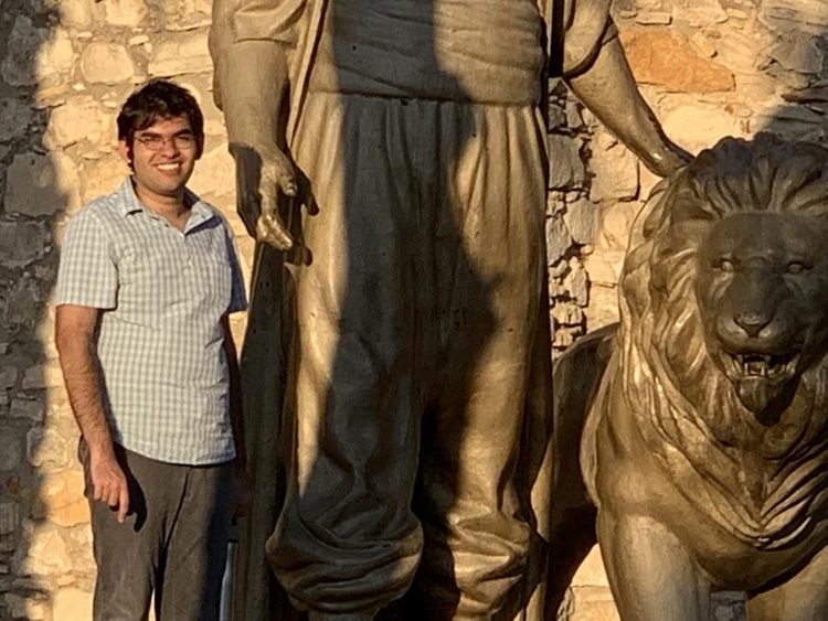 Arshan Hasan smiles as he stands next to an ancient statue of a giant petting a lion.