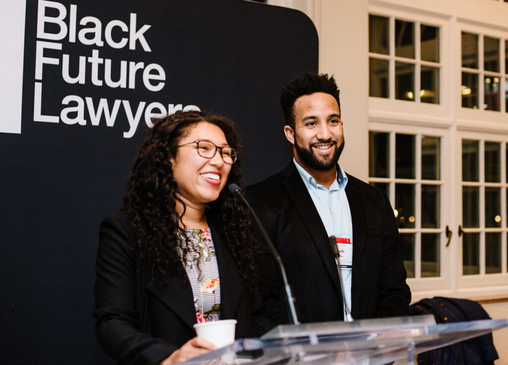 Rebecca Barclay Nguinambaye and Josh Lokko smile as they stand at a podium. A sign above them reads: Black Future Lawyers.