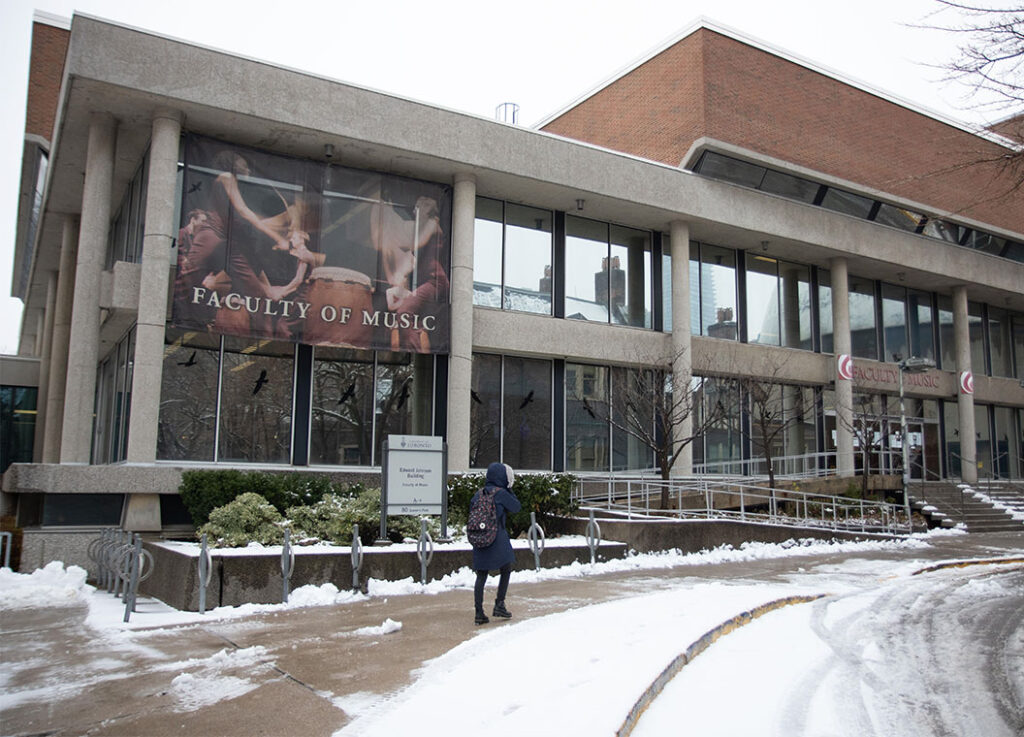 A woman walks along a snowy sidewalk in front of a building displaying a banner that reads Faculty of Music.