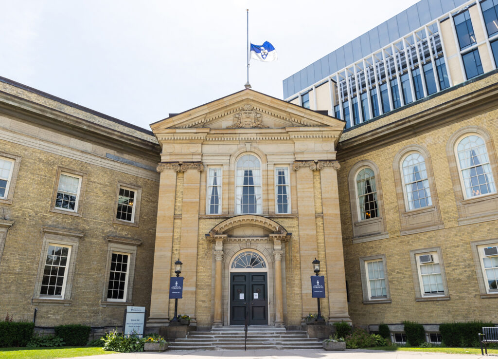 The University of Toronto flag flies at half mast over the roof of Simcoe Hall on a sunny day.