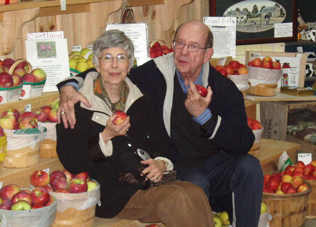 Cairine and Henderson Scott each hold an apple as they sit, arm in arm, on a shelf displaying baskets of apples.