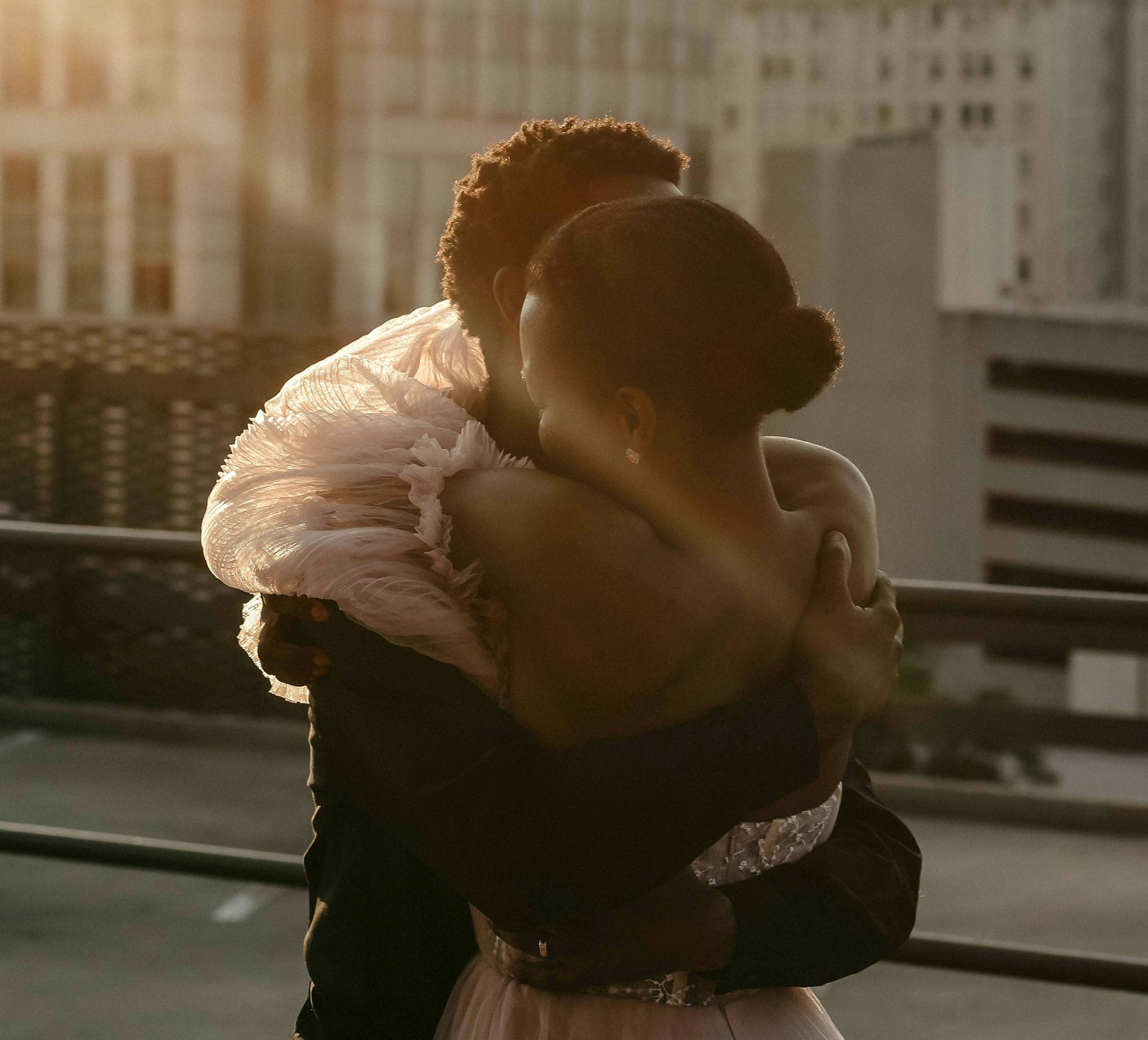 A young Black man and woman stand on a city balcony and hug each other tightly.
