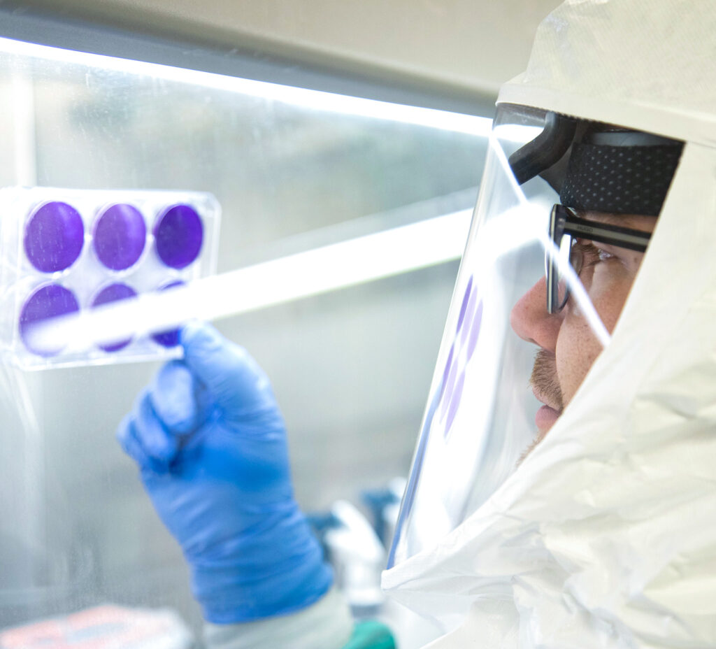 A man wears gloves and a full one-piece protective suit with helmet. He holds up and examines a petri dish with 6 samples.