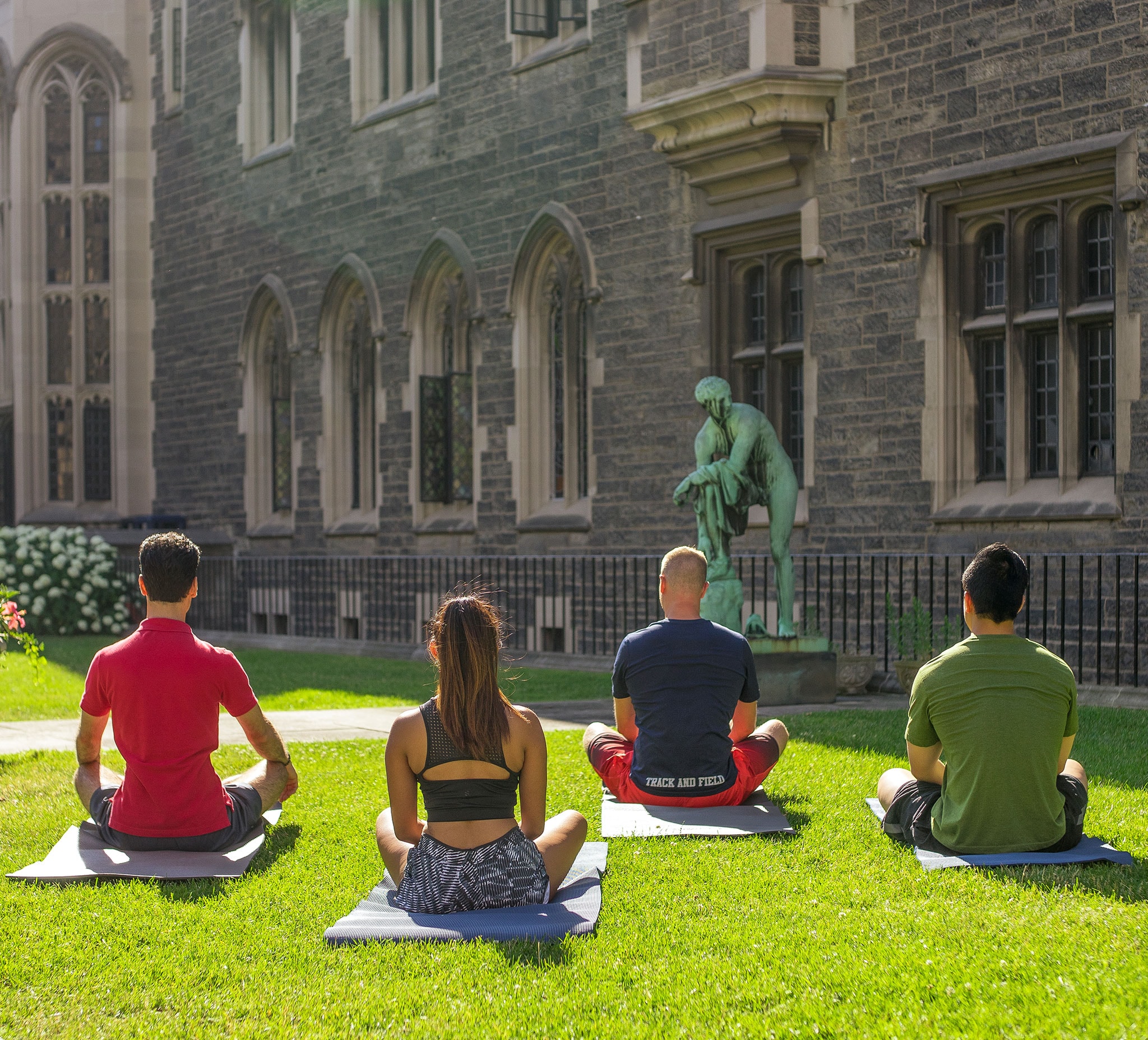 In the grassy courtyard of Hart House, on a sunny day, four people sit cross-legged on yoga mats.