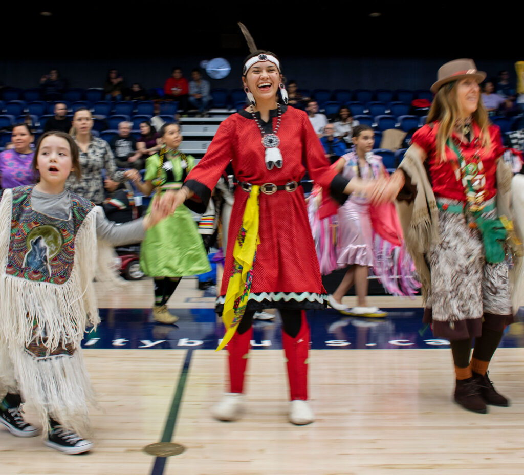 Indigenous women and girls wearing ceremonial regalia laugh as they dance, holding hands.