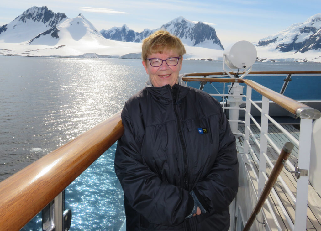 Joan McCalla smiles as she stands on the deck of a boat. Snowy mountains fill the shoreline.