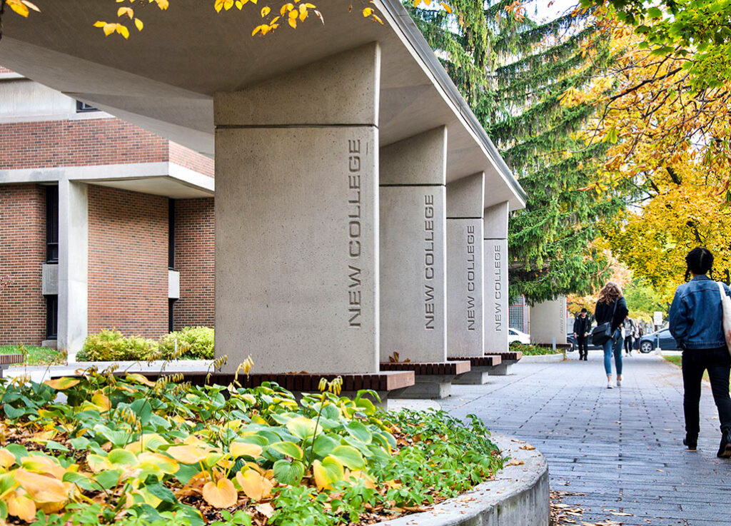 The words New College are engraved in concrete pillars holding up an inviting gateway with benches.