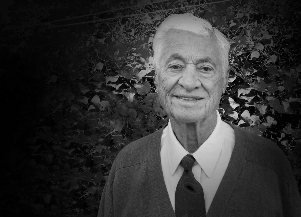 Nicolas Kordellas smiles quietly as he stands in front of a leafy hedge.