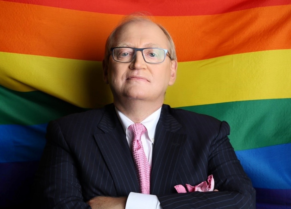 R. Douglas Elliott stands with arms folded in front of a rainbow flag.