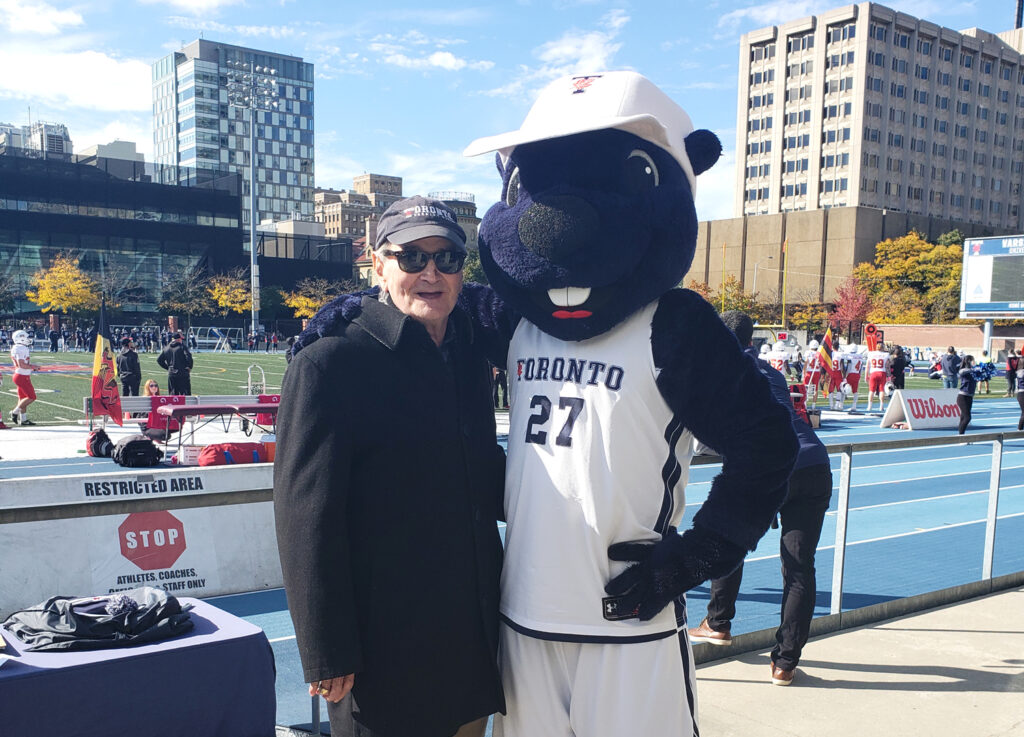 Ron Crawford smiles as he stands arm-in-arm with the True Blue beaver mascot on the sidelines of Varsity Stadium.