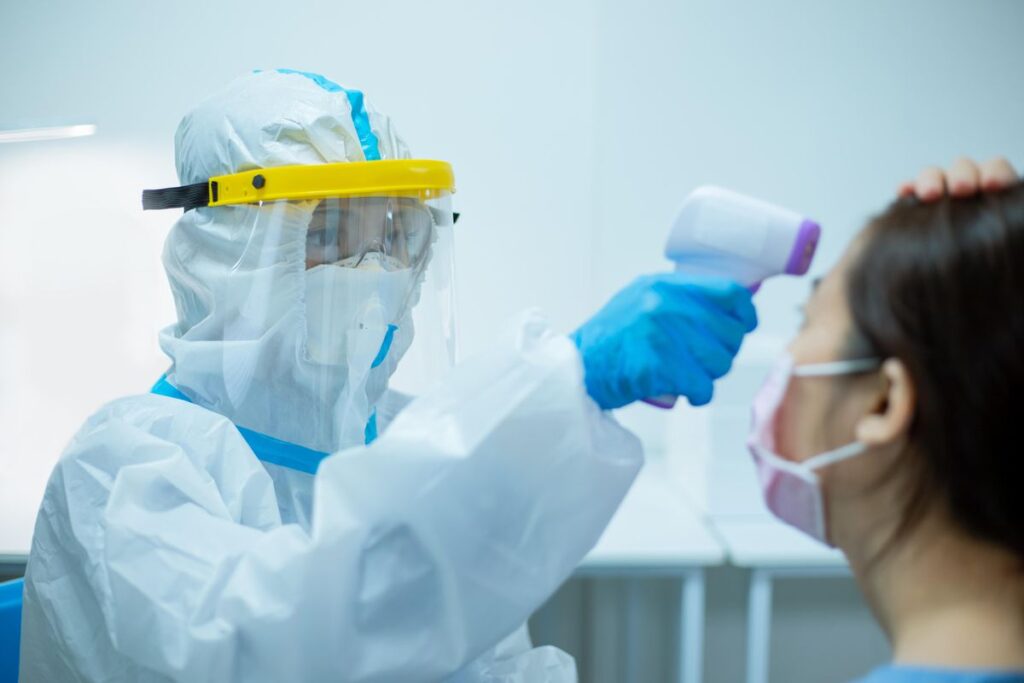 A worker in full protective suit, mask, face shield and gloves points a handheld device at the forehead of a masked patient.