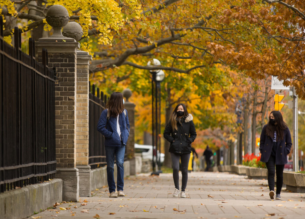 Three students in masks walk apart as they chat on a sidewalk under autumn trees.