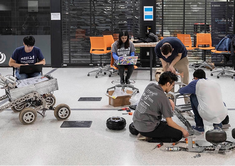 Five students crouch intently over their projects scattered across a classroom floor--a wheeled square object, a frame on tank tracks, and a drone.