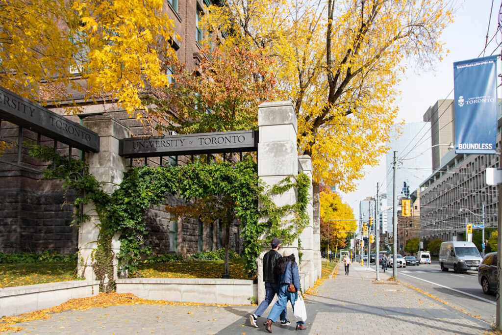 A couple walk out of the Alumni Gates on College Street, beneath the sign reading University of Toronto.