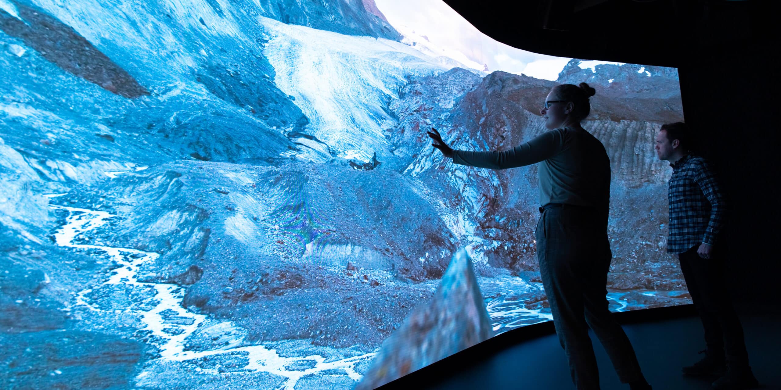 A woman reaches out to an enormous curved viewing screen in a dark room. The screen shows a mountain valley with a glacier.