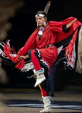 Miyopin Cheechoo dances, wearing traditional Indigenous regalia with moccasins, fringed shawl and a feather in her headband.