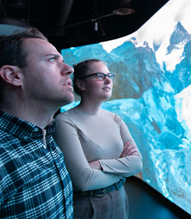 Two people look on in awe at a wall-sized screen that displays a mountain view.