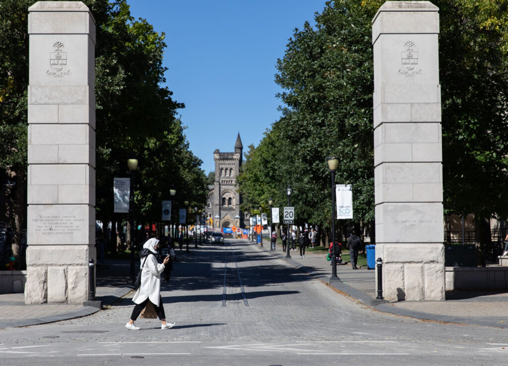A woman walks past two stone pillars flanking a cobbled street. University College can be seen at the end of the street.