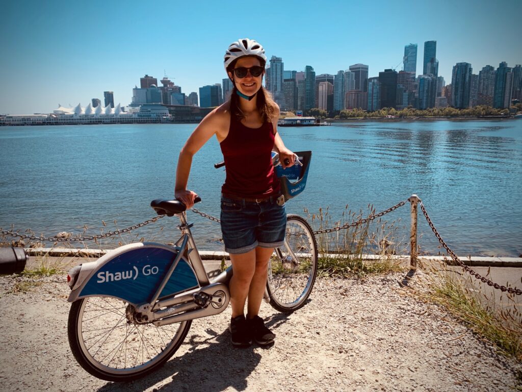 Carly MacEacheron smiles as she stands beside a ride-share bike, looking over a river to a busy city.